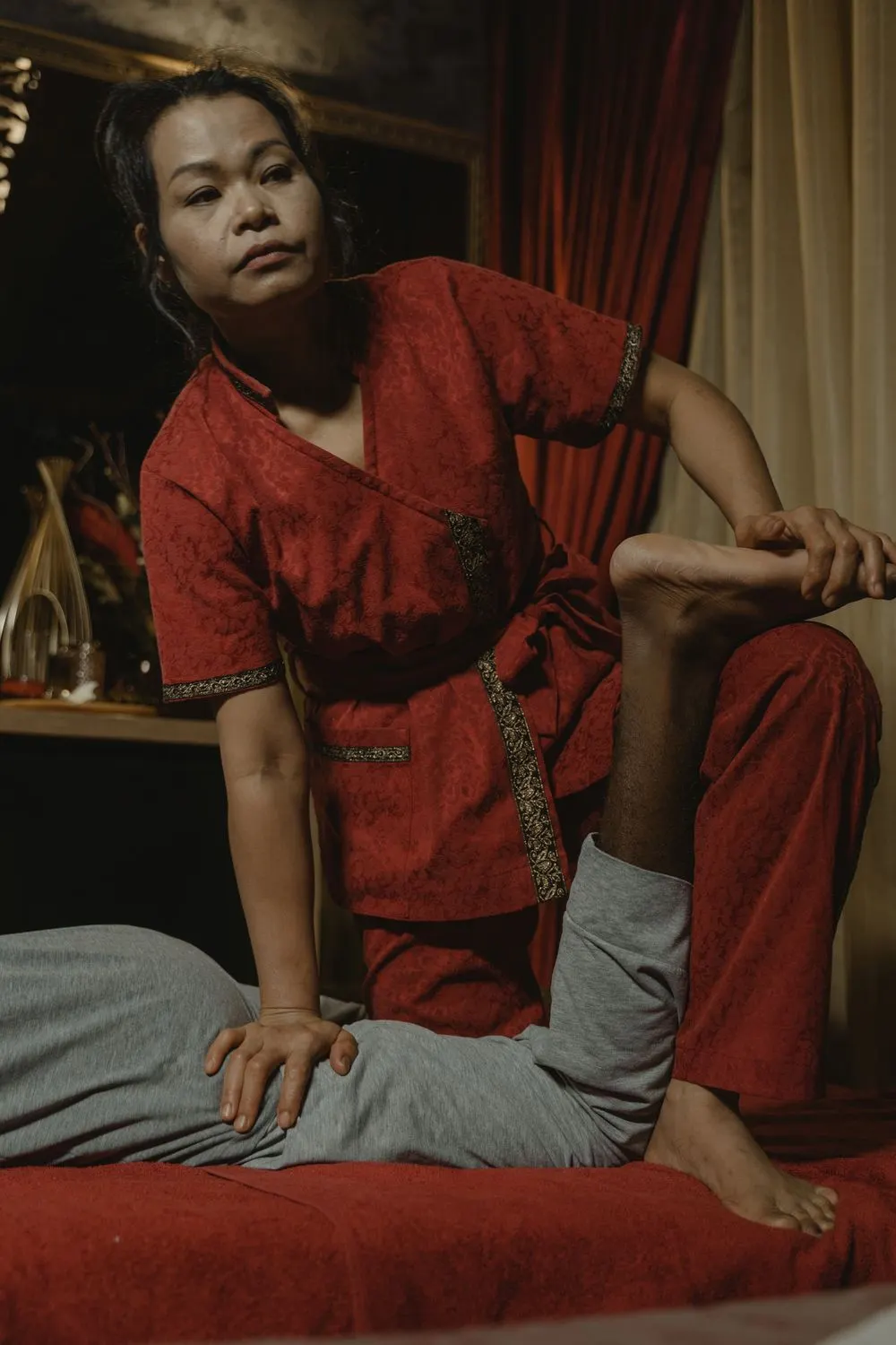 Therapist in traditional red outfit stretching a client's leg in a warm-lit room